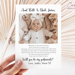 Puede incluir: Una tarjeta rectangular blanca con una fotografía de un bebé con un conjunto acanalado color crema y una diadema con un lazo grande. El texto dice: Aunt Kelli & Uncle James, un poema sobre padrinos y Will you be my godparents? Love, Ashley Marie.
