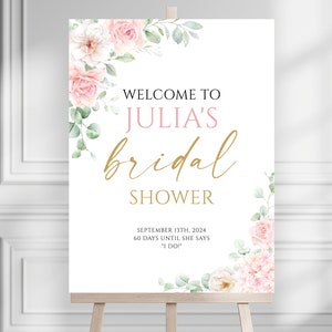 May include: A white sign on a wooden easel with floral accents, announcing "Welcome to Julia's Bridal Shower." The text is in black, pink, and gold, with the date September 13th, 2024, and a countdown.