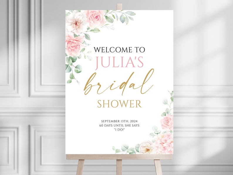 May include: A white sign on a wooden easel with floral accents, announcing "Welcome to Julia's Bridal Shower." The text is in black, pink, and gold, with the date September 13th, 2024, and a countdown.