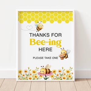 May include: A framed print with a white border features a yellow honeycomb pattern at the top. The text reads "THANKS FOR Bee-ing HERE" with bee illustrations. A floral border of daisies and bees is at the bottom. "PLEASE TAKE ONE" is written at the bottom.
