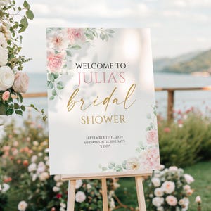May include: A sign on an easel, announcing "Welcome to Julia's bridal shower". The sign features pink and gold lettering, floral decorations, and the date September 13th, 2024. The scene is set outdoors with flowers and a lake in the background.