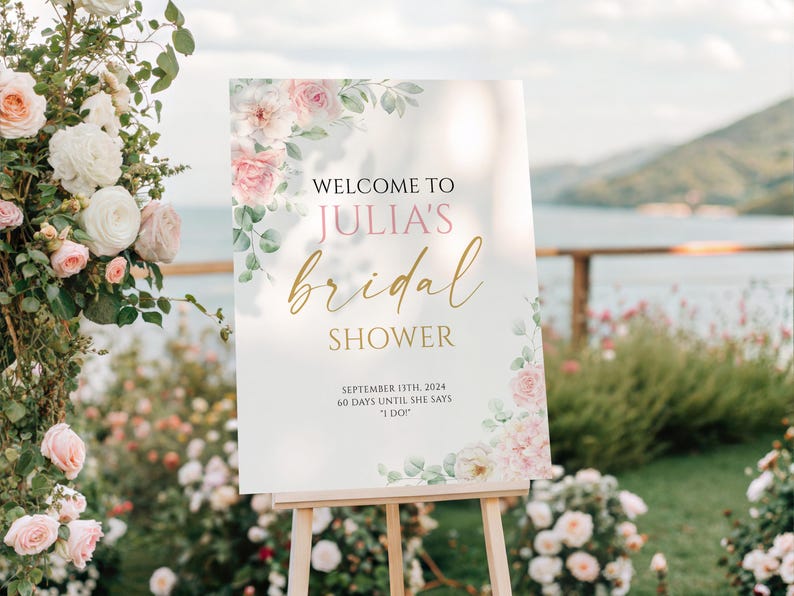 May include: A sign on an easel, announcing "Welcome to Julia's bridal shower". The sign features pink and gold lettering, floral decorations, and the date September 13th, 2024. The scene is set outdoors with flowers and a lake in the background.