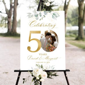 May include: A white sign on a black easel, celebrating a 50th anniversary. The sign features gold script that reads "Celebrating 50 Years" and the names "David & Margaret" with a photo of a couple. Floral accents and the date February 25, 2026 are also included.