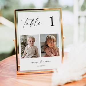 Op de afbeelding: Een goudomrand tafelnummerkaartje met het woord "Table 1" in elegant schrift. De kaart bevat twee foto's van kinderen, met namen en een datum eronder. De kaart wordt weergegeven op een houten oppervlak.