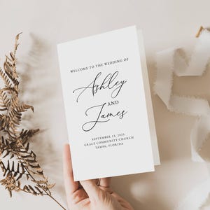Könnte beinhalten: Ein weißes Hochzeitsprogramm mit den Namen "Ashley and James" in eleganter Schrift. Das Programm enthält das Hochzeitsdatum und den Ort. Eine Hand hält das Programm, mit einem getrockneten Farn und weißem Band in der Nähe.