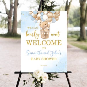Op de afbeelding: Een welkomstbord voor een babyshower op een zwart schildersezel. Het bord toont een teddybeer in een luchtballon gemaakt van gouden, witte en blauwe ballonnen. De tekst luidt "We can bearly wait WELCOME to Samantha and John's BABY SHOWER".