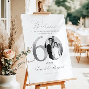 May include: A white rectangular sign on a wooden easel featuring the text Welcome to the 60th wedding anniversary celebration of, a large silver number 60, a circular black and white wedding photo, the names David & Margaret, and February 25, 2025.