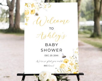 May include: A white baby shower welcome sign on a black easel. The sign has gold script that reads "Welcome to Ashley's Baby Shower" with the date. Yellow floral and bee illustrations decorate the sign.
