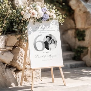 May include: A white rectangular wedding anniversary sign on a wooden easel featuring the text Welcome to the 60th wedding anniversary celebration of, the number 60, a black and white photo of a couple, the names David & Margaret, and February 25, 2023.