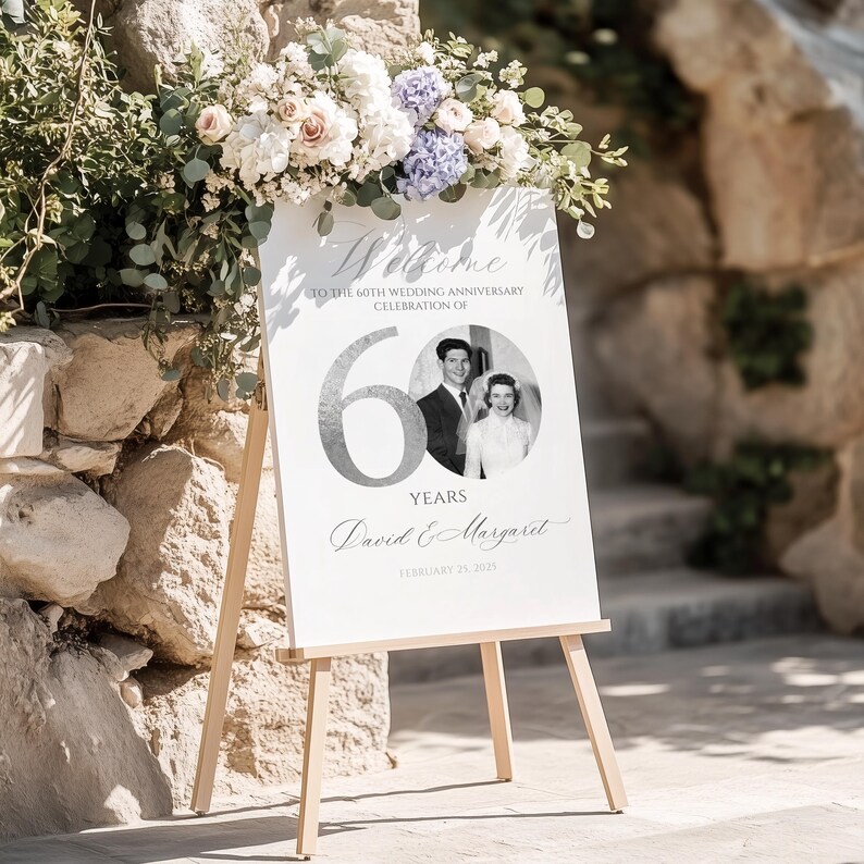 May include: A white rectangular wedding anniversary sign on a wooden easel featuring the text Welcome to the 60th wedding anniversary celebration of, the number 60, a black and white photo of a couple, the names David & Margaret, and February 25, 2023.