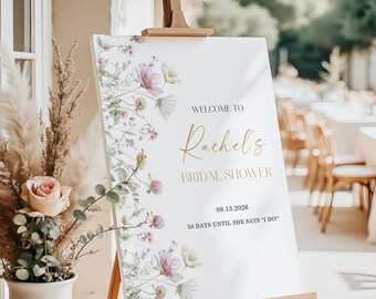 May include: A white sign on a wooden easel with floral accents and gold text that reads "WELCOME TO Rachel's BRIDAL SHOWER 09.13.2026 58 DAYS UNTIL SHE SAYS 'I DO!'" The sign is decorated with purple and white flowers.