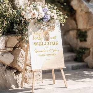 May include: A welcome sign for a baby shower, featuring the text "We can bearly wait WELCOME to Samantha and John's BABY SHOWER." The sign has a teddy bear in a basket and is decorated with gold and beige tones. It is displayed on a wooden easel.