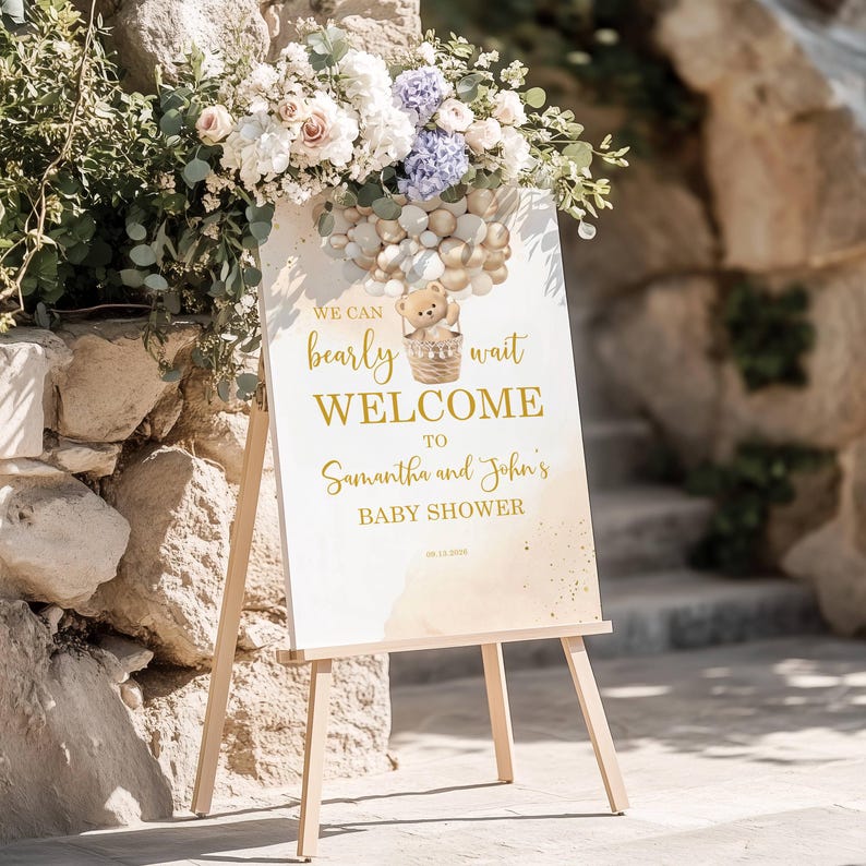 May include: A welcome sign for a baby shower, featuring the text "We can bearly wait WELCOME to Samantha and John's BABY SHOWER." The sign has a teddy bear in a basket and is decorated with gold and beige tones. It is displayed on a wooden easel.