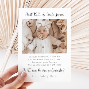 Pode incluir: Um cartão branco com uma fotografia de um bebé com uma fita de laço e uma chupeta. O cartão diz "Tia Kelli e Tio James" e "Queres ser meus padrinhos?" com um poema. O cartão é segurado em frente a um fundo bege.