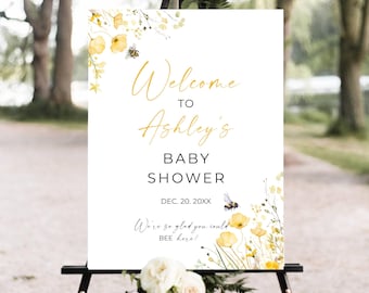 May include: A white sign on a black easel with the text "Welcome to Ashley's BABY SHOWER" in gold script. The sign includes floral and bee illustrations, with the phrase "We're so glad you could BEE here!"