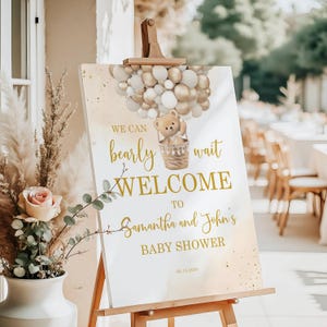 May include: A baby shower welcome sign on an easel. The sign features gold text that reads "We can bearly wait to welcome to Samantha and John's Baby Shower." A teddy bear is in a basket attached to balloons.