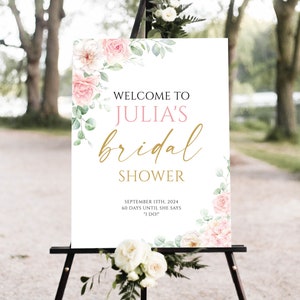 May include: A white sign on a black easel with the text "WELCOME TO JULIA'S bridal SHOWER". The sign is decorated with pink roses and green leaves. The date "SEPTEMBER 13TH, 2024" is also displayed.