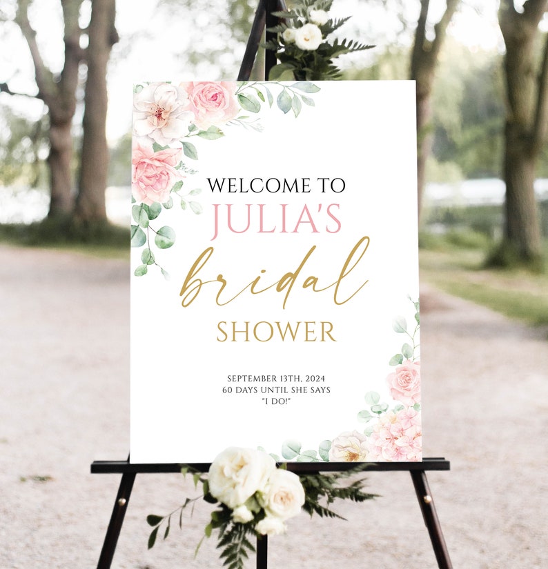 May include: A white sign on a black easel with the text "WELCOME TO JULIA'S bridal SHOWER". The sign is decorated with pink roses and green leaves. The date "SEPTEMBER 13TH, 2024" is also displayed.