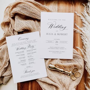 Op de afbeelding: Trouwuitnodigingsset met twee witte kaarten met elegante kalligrafie. Eén kaart leest "Welkom bij de bruiloft van Julia & Robert" met datum en tijd. Een gouden lakzegel en een houten plank maken de scène compleet.