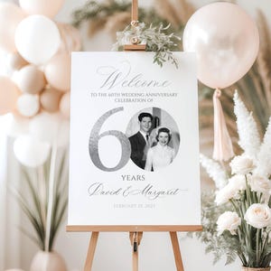May include: A white rectangular sign on a wooden easel featuring the text Welcome to the 60th wedding anniversary celebration of, a large silver number 60, a circular black and white photo of a couple, the names David & Margaret, and February 25, 2025.
