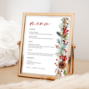 May include: A rectangular gold metal frame holding a printed menu card. The menu features the word menu in red script, followed by black text listing starters, mains, and desserts. A watercolor illustration of pine branches, red berries, and a pinecone.