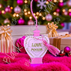 May include: A pink heart-shaped ornament with the words "LOVE POTION" on a banner. The ornament is hanging with a pink string and tassel. The text "Digital File - SVG" and "Not a physical item" are also visible. The background includes Christmas ornaments and gifts.