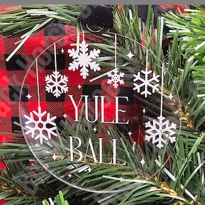May include: A clear, circular ornament with white snowflake designs and the words "YULE BALL" in white. The ornament hangs against a backdrop of red and black plaid fabric and green garland. The text "Digital File *SVG*" and "Not a Physical Item" are also present.