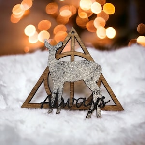 May include: A wooden ornament shaped like a triangle with a circle inside. The triangle has a deer silhouette inside and the word "Always" written below. The ornament is covered in silver glitter.