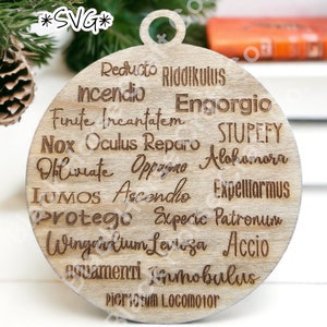 May include: A round wooden ornament with a light brown wood grain. The ornament is decorated with a list of spells from the Harry Potter series, including "Reducto", "Riddikulus", "Incendio", "Engorgio", "Finite Incantatem", "Nox", "Oculus Reparo", "Stupefy", "Diluviate", "Oppaiono", "Alahainora", "Lumos", "Ascendio", "Expelliarmus", "Protego", "Expecto Patronum", "Wingardium Leviosa", "Accio", "Aguamenti", "Immobulus", and "Piertotum Locomotor".