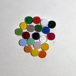 May include: Assortment of small, round glass tiles in various colors, including red, green, blue, yellow, white, and black. The tiles are arranged in a circular pattern on a white surface.