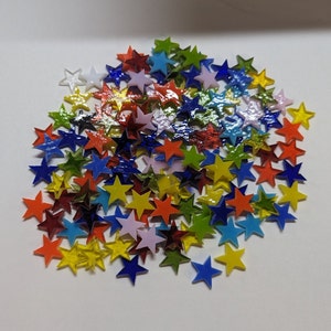 May include: A collection of colorful star-shaped glass pieces in various colors, including red, yellow, blue, green, and purple. The stars are scattered on a white surface, creating a vibrant and playful display. These decorative stars are ideal for crafts.