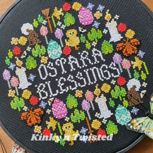 May include: A black embroidery hoop with a colorful cross-stitch design featuring the text "Ostara Blessings". The design includes flowers, eggs, butterflies, candles, and other spring-themed imagery.