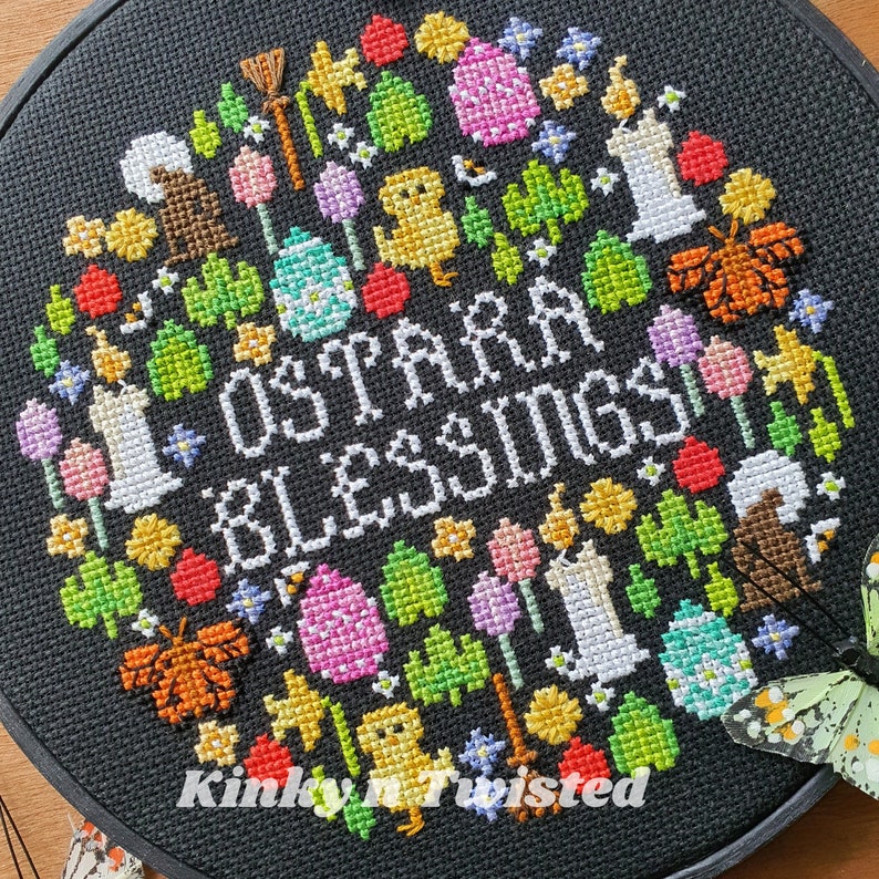 May include: A black embroidery hoop with a colorful cross-stitch design featuring the text "Ostara Blessings". The design includes flowers, eggs, butterflies, candles, and other spring-themed imagery.