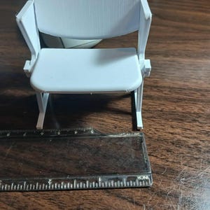 May include: A small, light gray miniature chair with a seat, back, and armrests. The chair is placed on a wooden surface next to a ruler. The chair measures approximately 3 inches in height.