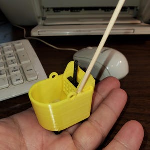 May include: A miniature yellow bucket with a black mop and a wooden handle. The bucket has a handle on each side and sits on two small black wheels. The miniature cleaning supplies are held in a hand.