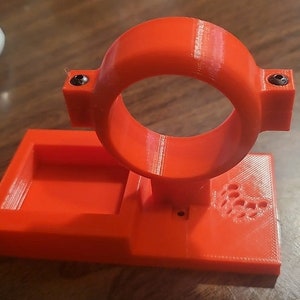 May include: A bright orange 3D printed stand with a circular opening and a rectangular base. The stand has a small hole on the side and a pattern of small circles on the base.