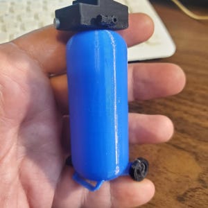 May include: A miniature blue air compressor model with black details, including a top component and small wheels. The model is held in a hand, showcasing its size and design.