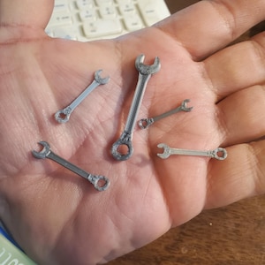 May include: A collection of small, silver-colored wrenches of varying sizes. The wrenches are arranged in a hand, showcasing their miniature design and metallic finish. The tools are likely for a model or miniature project.