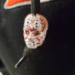 May include: A white hockey mask with red paint splatters and black holes. The mask is attached to a black cord.