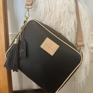 May include: Black crossbody bag with a beige trim and strap. The bag features a gold zipper and hardware, a beige patch with the brand name, and a black tassel. The bag is hanging against a white fluffy background.
