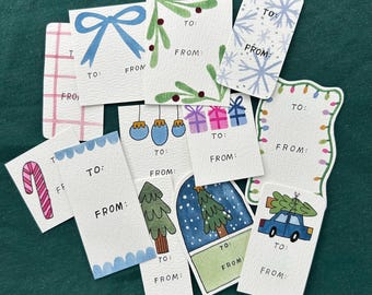 May include: A collection of hand-painted gift tags with various holiday-themed illustrations. Designs include a blue bow, snowflakes, Christmas lights, ornaments, presents, a candy cane, a Christmas tree, and a car carrying a tree. Each tag has space for "To:" and "From:"