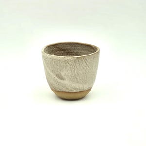 May include: A ceramic cup with a textured, off-white exterior and a brown rim. The base of the cup is a natural tan colour. The cup has a simple, rounded shape, suitable for beverages.