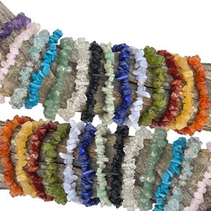 May include: A selection of gemstone chip bracelets in a variety of colours, such as purple, pink, blue, green, clear, black, and orange. The bracelets are strung on elastic and displayed on a wooden surface.