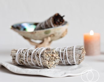May include: Two white sage smudge sticks tied with white string on a white plate. The smudge sticks are in focus, while a seashell and a candle are blurred in the background.