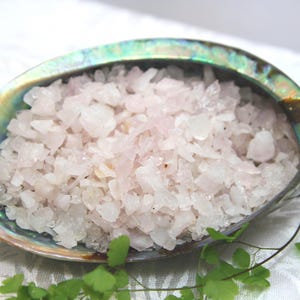 May include: A close-up shot of a natural abalone shell filled with pale pink, crystalline salt. The shell displays iridescent green, blue, and gold colours. Green foliage is visible in the foreground, adding a natural touch.
