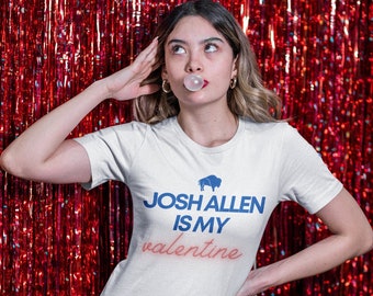 May include: A white t-shirt with blue text that reads "Josh Allen is my valentine" and a small blue buffalo silhouette above the text. The person wearing the shirt is standing in front of a red sparkly background.