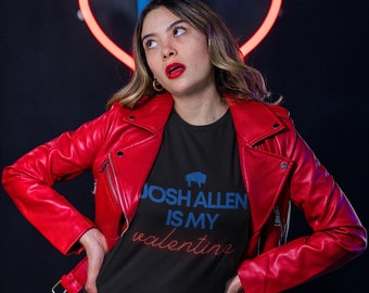May include: A woman wearing a red leather jacket over a black t-shirt with the text "Josh Allen is my valentine" and a blue buffalo silhouette. She is standing in front of a neon pink heart.