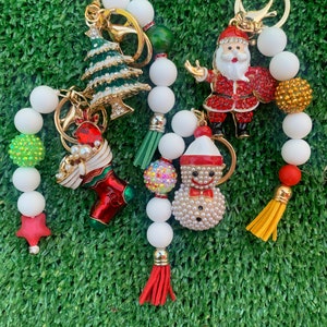 May include: A set of Christmas-themed keychains with white beads, red, green, and gold accents. The keychains feature a Christmas tree, Santa Claus, a stocking, a snowman, and a star.