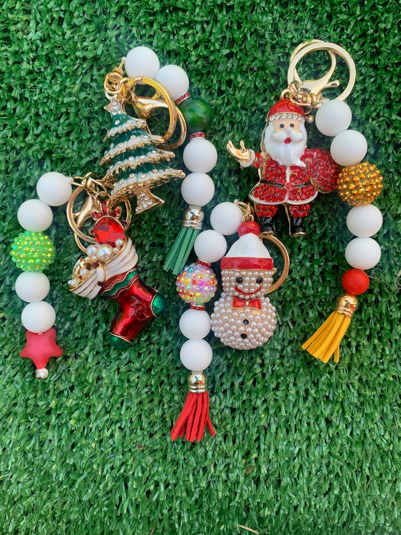 May include: A set of Christmas-themed keychains with white beads, red, green, and gold accents. The keychains feature a Christmas tree, Santa Claus, a stocking, a snowman, and a star.