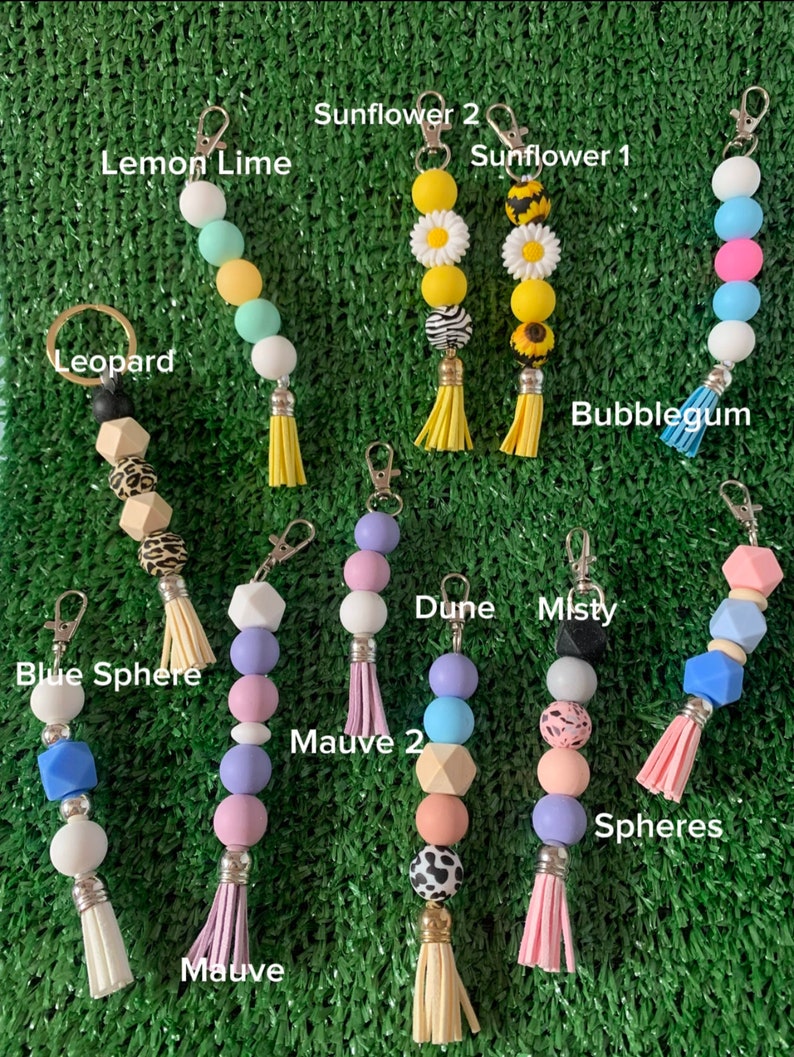 May include: Assortment of colorful beaded keychains with tassels.  The keychains feature various designs, including a sunflower, a leopard print, a blue sphere, and a mauve color scheme.  The keychains are labeled with names such as "Sunflower 1", "Lemon Lime", "Bubblegum", "Dune", "Misty", "Mauve", "Mauve 2", and "Spheres".